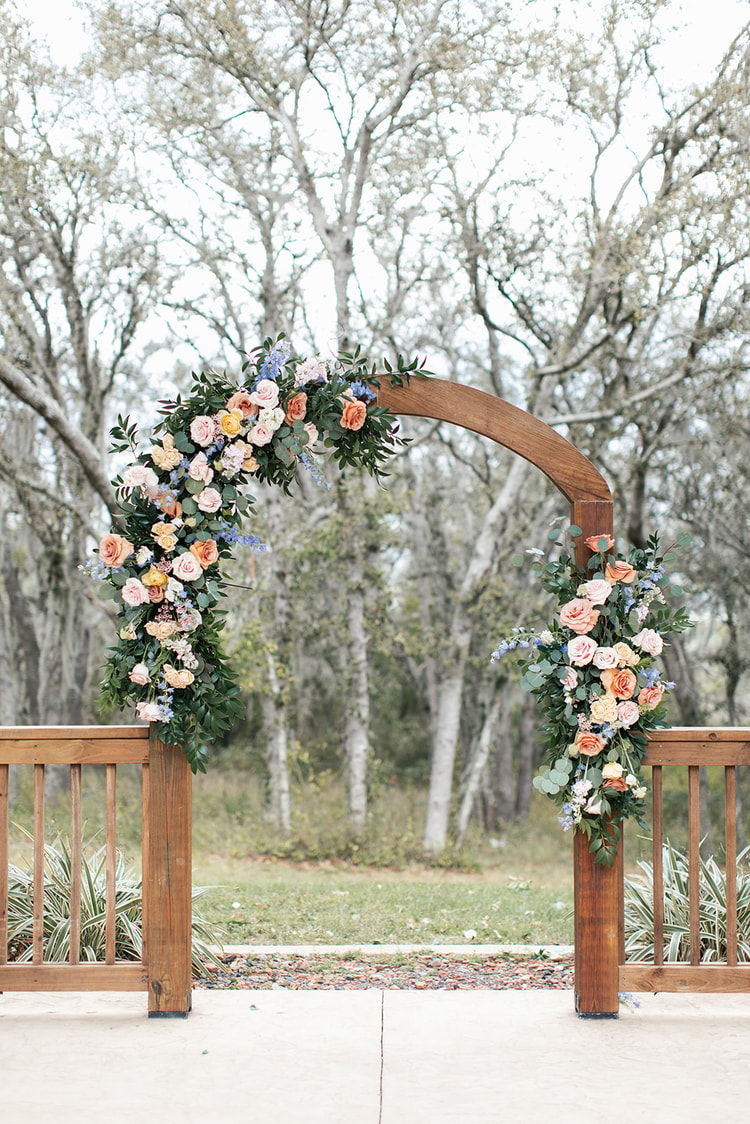 Wooden ceremony arch draped with peach and blush roses and blue delphinium, set against bare winter trees at Sterling Event Venue.