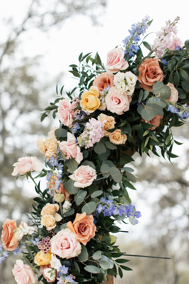 Close-up of a lush ceremony arch garland with pink, peach, and yellow roses, blue delphinium, and eucalyptus in soft daylight.