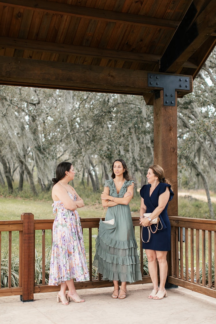 Three wedding guests in floral, sage, and navy dresses chat under a wooden pavilion with oak trees in the background.