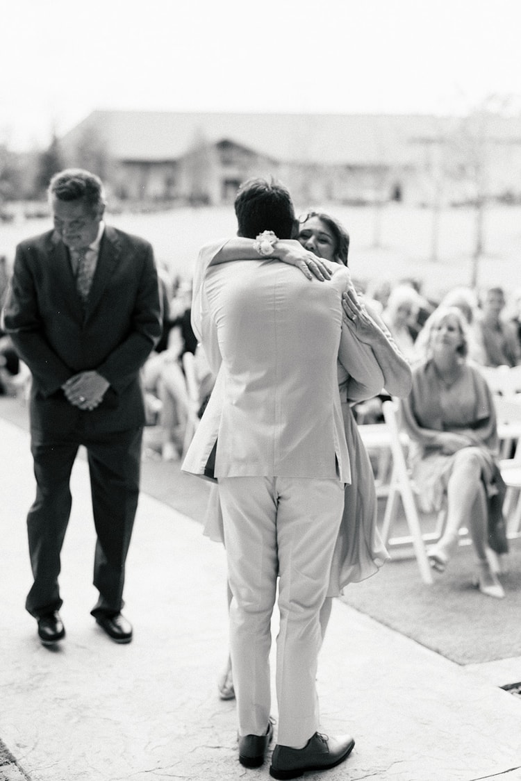 Black and white photo of the groom hugging a guest before the ceremony while another man in a suit looks on.