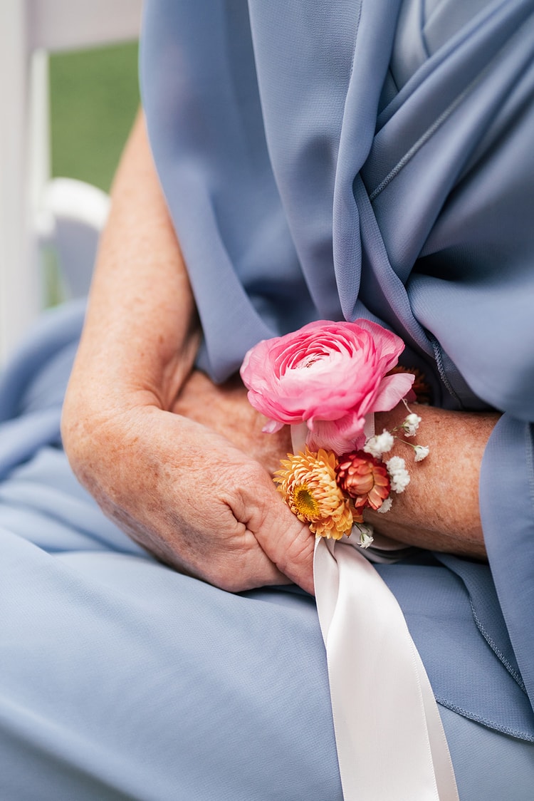 Close-up of an older woman's hands resting in her lap, adorned with a pink and orange floral wrist corsage tied in ribbon.