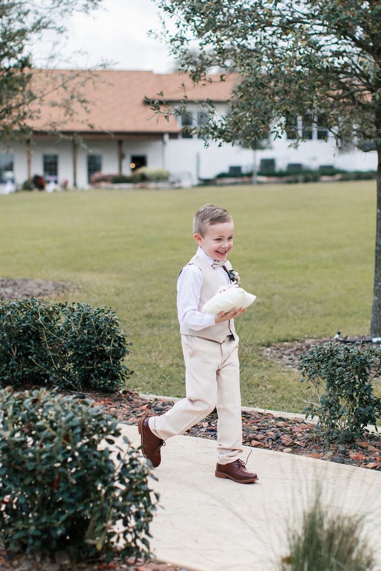 Ring bearer in a tan vest and bow tie smiles while carrying a ring pillow across a garden path at the wedding venue.