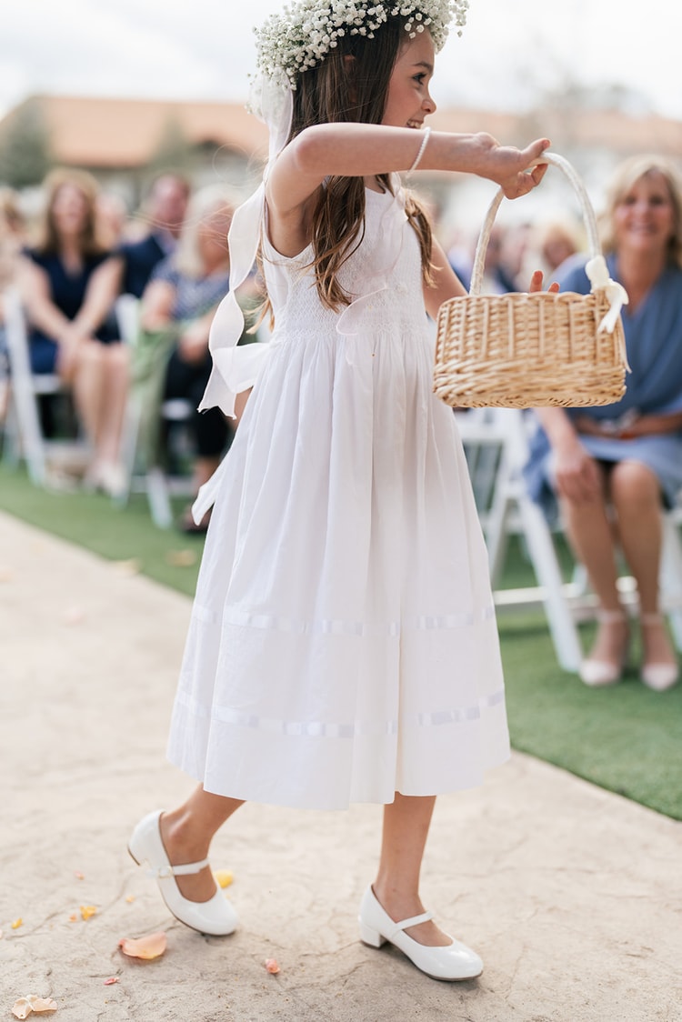 Flower girl in a white dress and baby's breath crown carries a woven basket down the aisle, trailing rose petals.