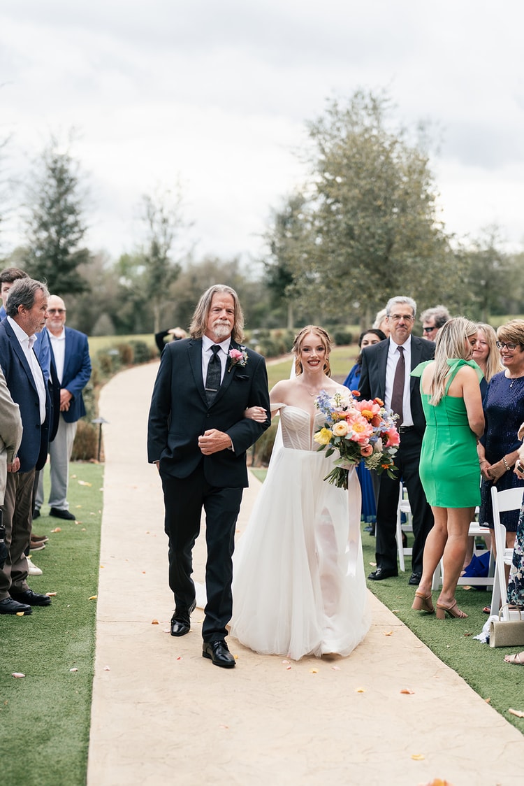 Bride walks down the aisle on her father's arm, smiling and holding a bright bouquet of coral, yellow, and blue flowers.