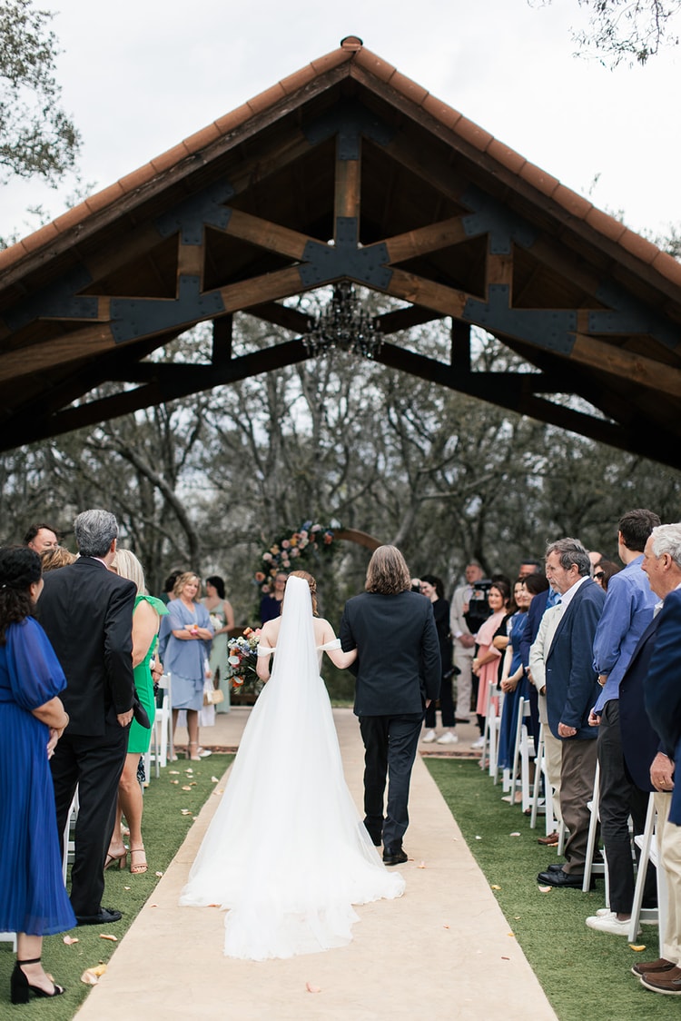 Bride and her father walk arm in arm down the aisle beneath a timber-framed pavilion at Sterling Event Venue as guests stand and watch.