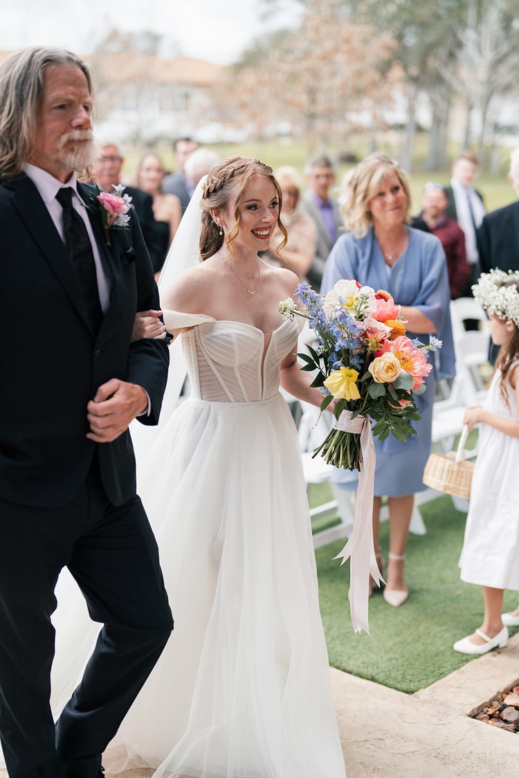Bride in an off-shoulder gown and veil smiles while walking down the aisle arm-in-arm with her father, colorful bouquet in hand.