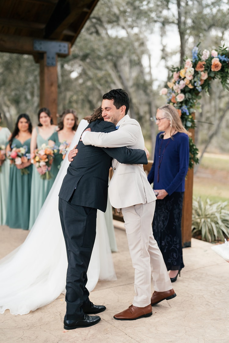 Groom embraces a family member on the ceremony stage as bridesmaids in sage green hold bouquets in the background.