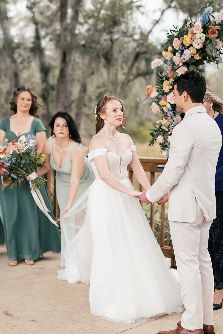 Bride and groom hold hands and gaze at each other during their vows beneath a floral arch, bridesmaids looking on.