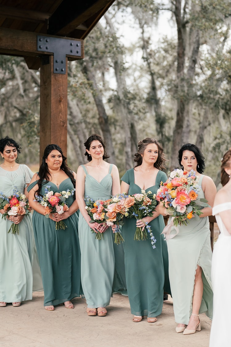 Bridesmaids in sage and dusty green dresses stand together holding colorful bouquets of coral roses and blue delphinium under the pavilion.