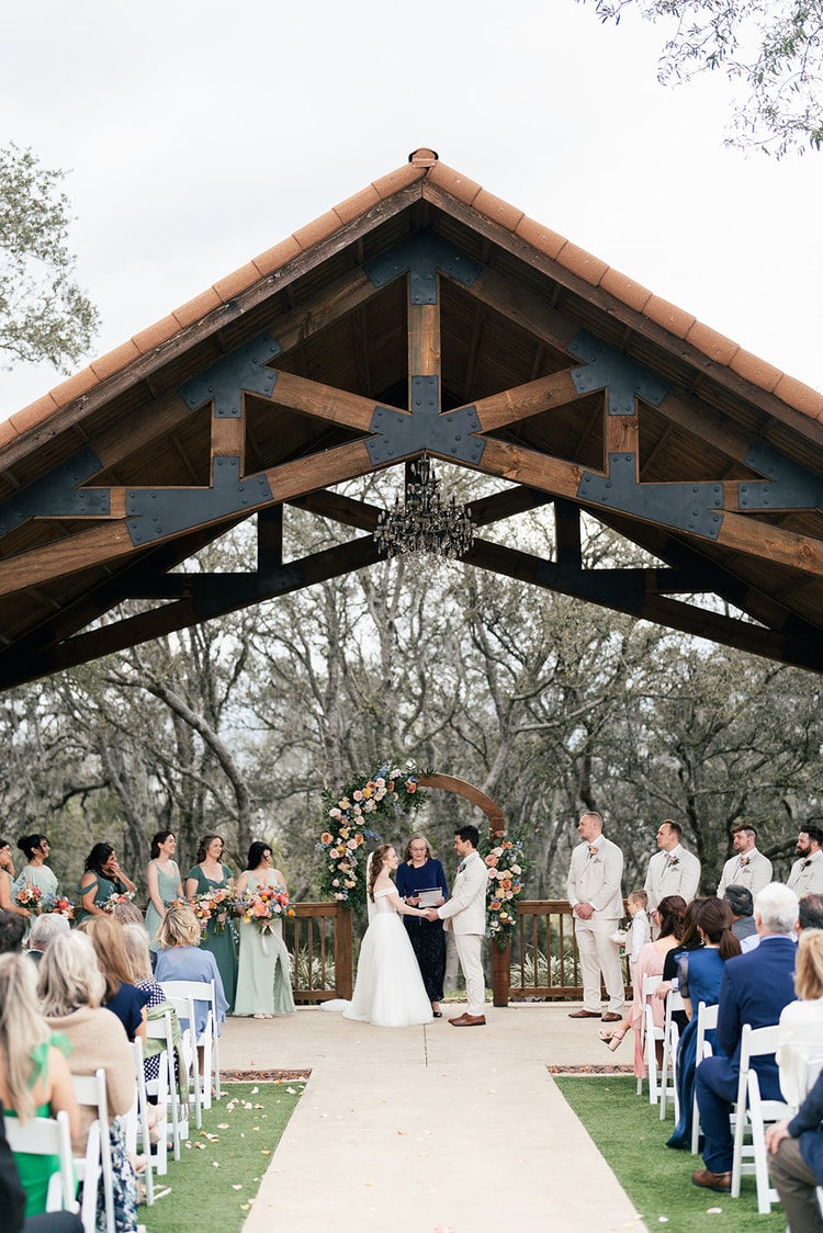 Bride and groom hold hands at the altar surrounded by bridesmaids and groomsmen as the officiant reads from her notes.