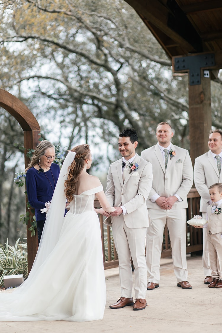 Bride faces her groom and groomsmen at the altar, holding hands as the ceremony begins under the timber pavilion.
