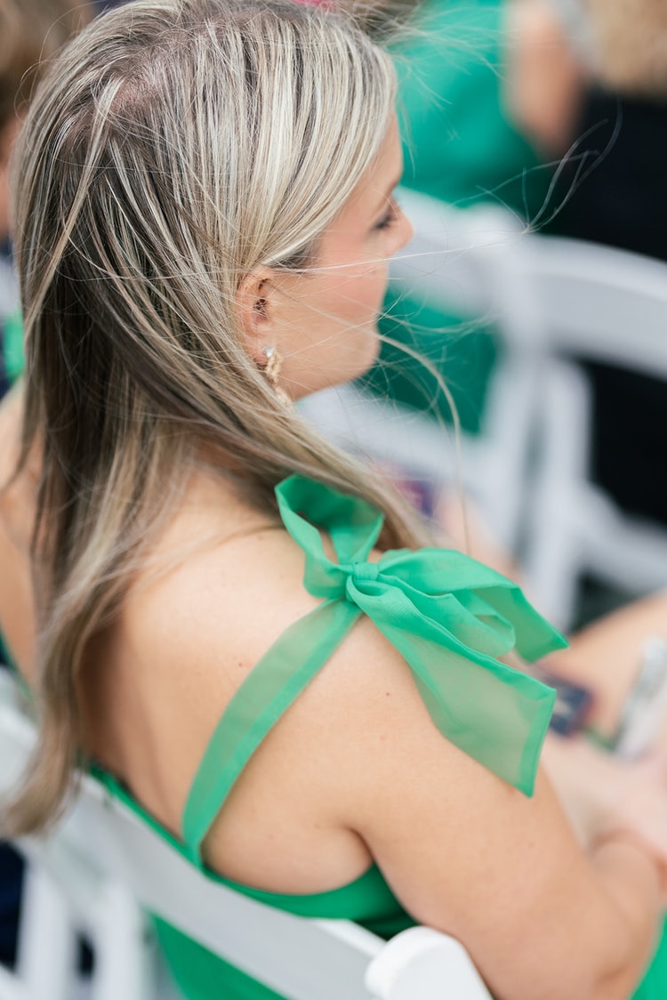 Close-up of a wedding guest's shoulder with a green chiffon bow, hair blowing in the wind during the outdoor ceremony.