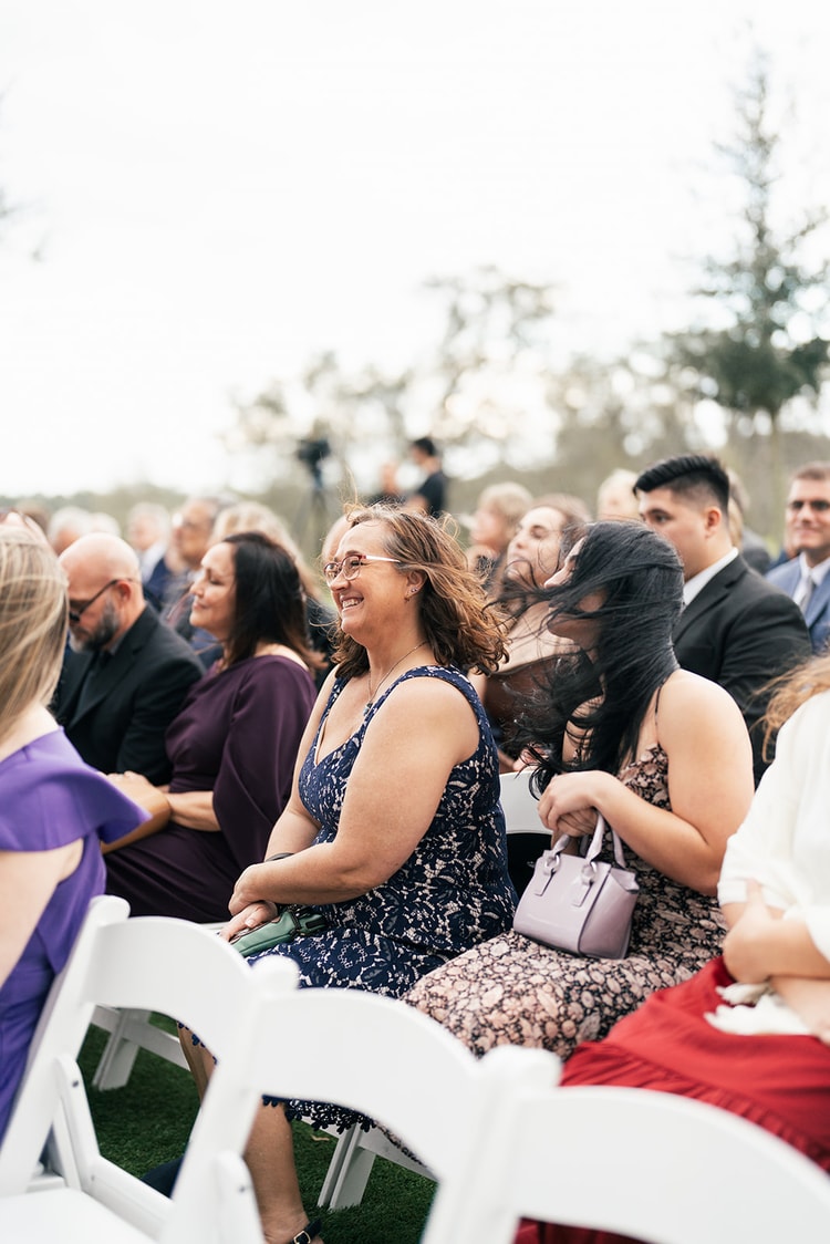 Wedding guest in a navy lace dress smiles from her seat as the wind blows through the crowd during the outdoor ceremony.