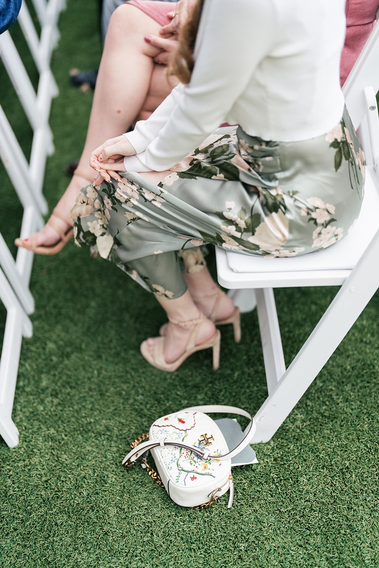 Detail shot of a seated guest in a floral dress with nude heels and a white embroidered handbag on the artificial turf lawn.