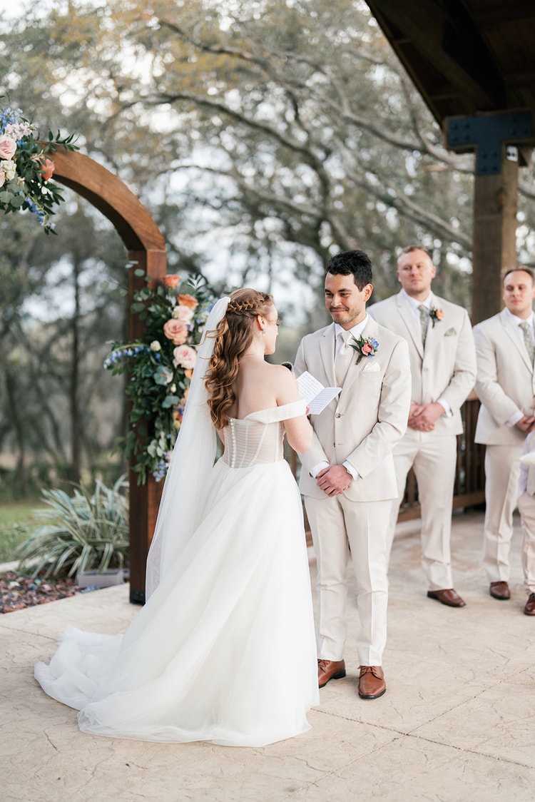Bride reads her vows aloud to the groom from a small notecard as groomsmen in tan suits stand behind him.