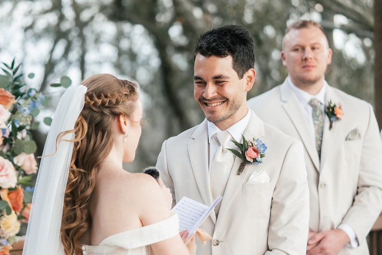 Groom smiles while listening to the bride's vows, groomsman looking on softly in the background under the pavilion.