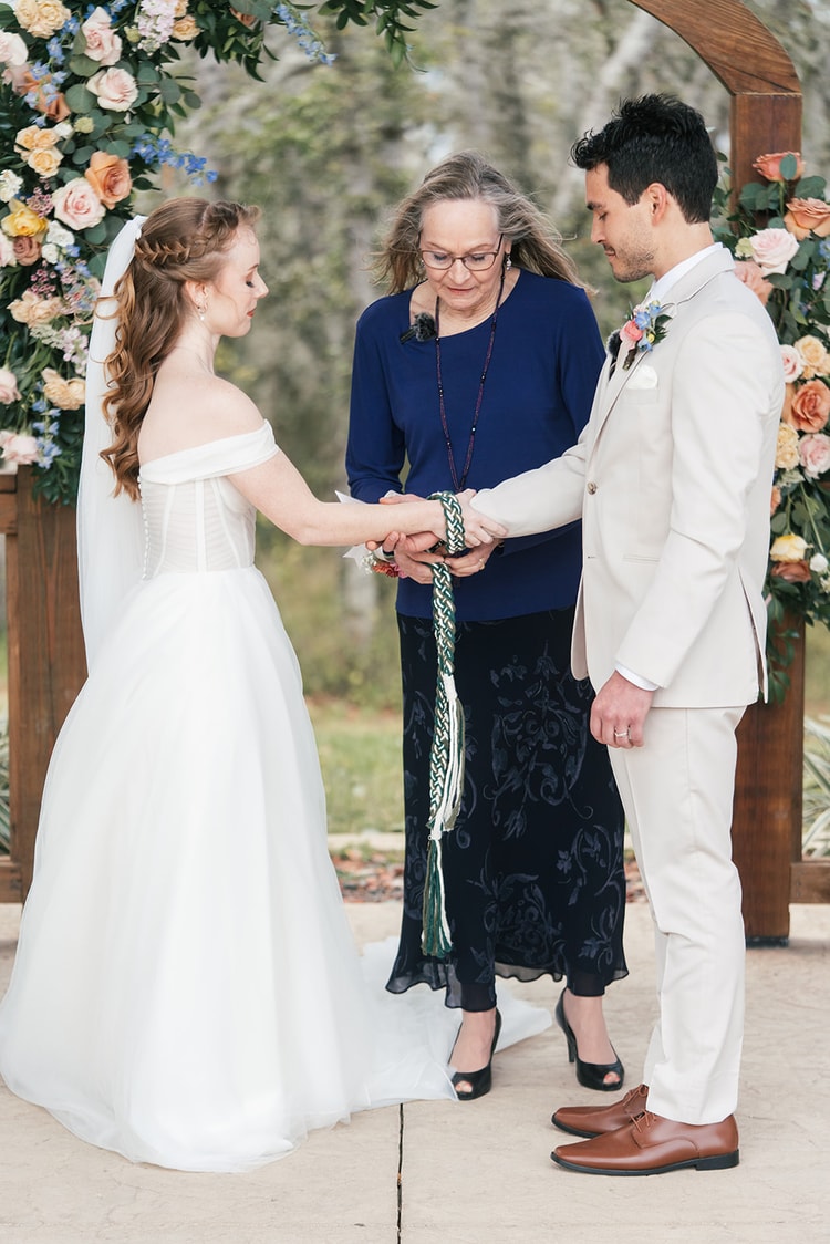 Bride and groom hold a handfasting cord while the officiant in a navy top guides the ritual during their vows.