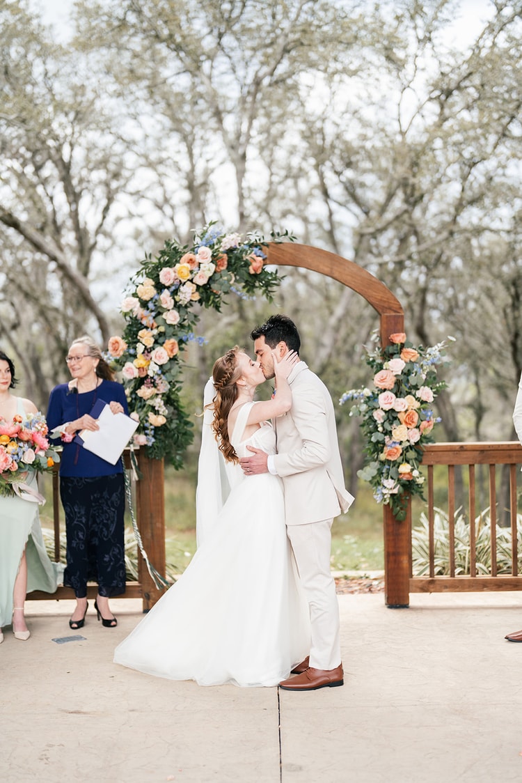 Bride and groom share their first kiss as a married couple beneath the flower-covered wooden arch.