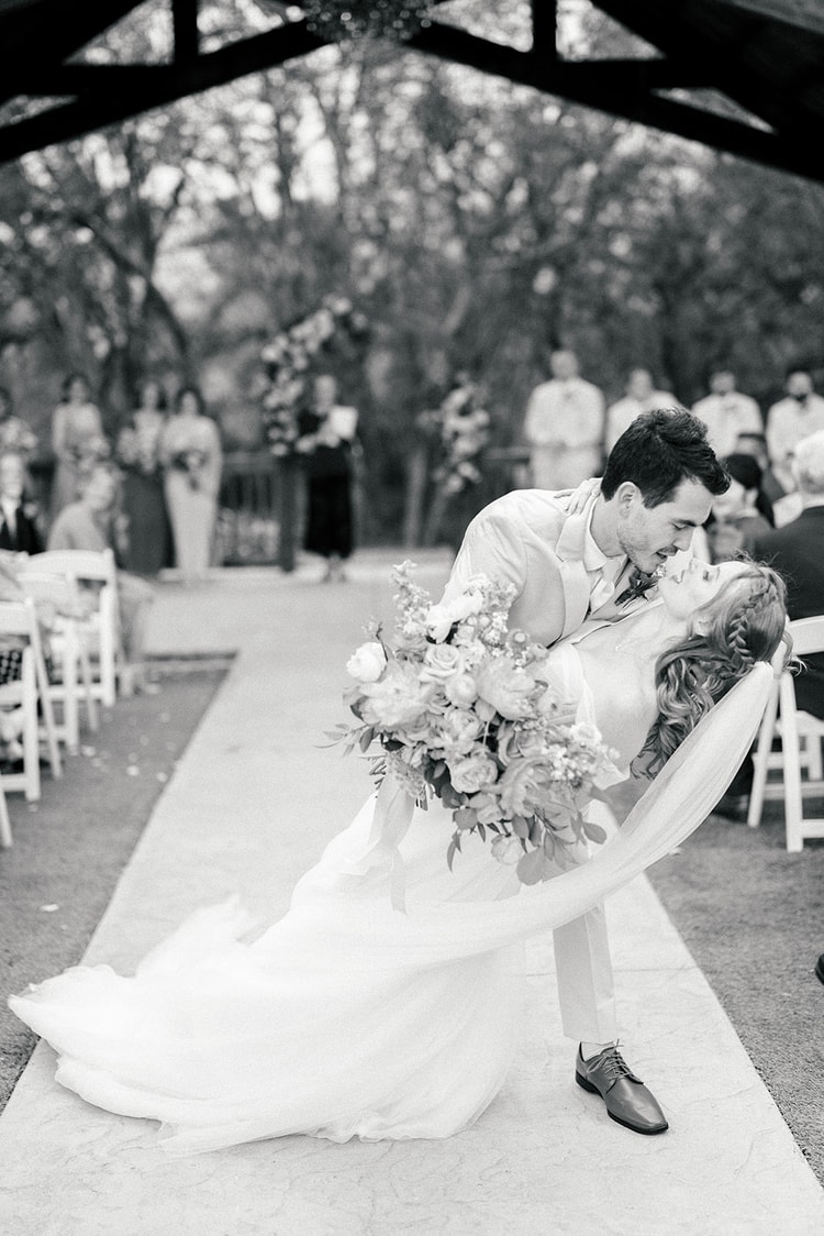Black and white photo of the groom dipping his bride for a kiss on the aisle after their ceremony, bouquet in hand.