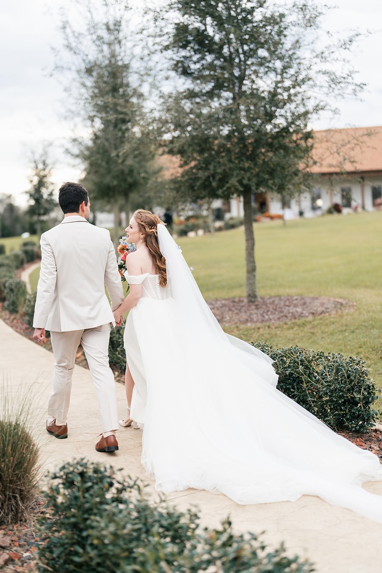 Bride and groom walk hand-in-hand along a garden path near the venue's clubhouse, her veil trailing behind her.