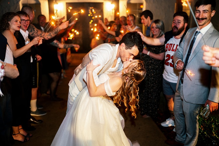 Bride and groom kiss during their sparkler send-off outside the venue at night, guests cheering on both sides.