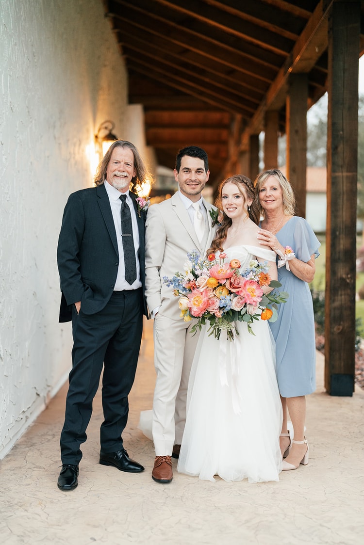 Bride and groom pose with her parents under a wooden-beamed walkway at Sterling Event Venue, the bride holding a large bouquet of coral and blue flowers.