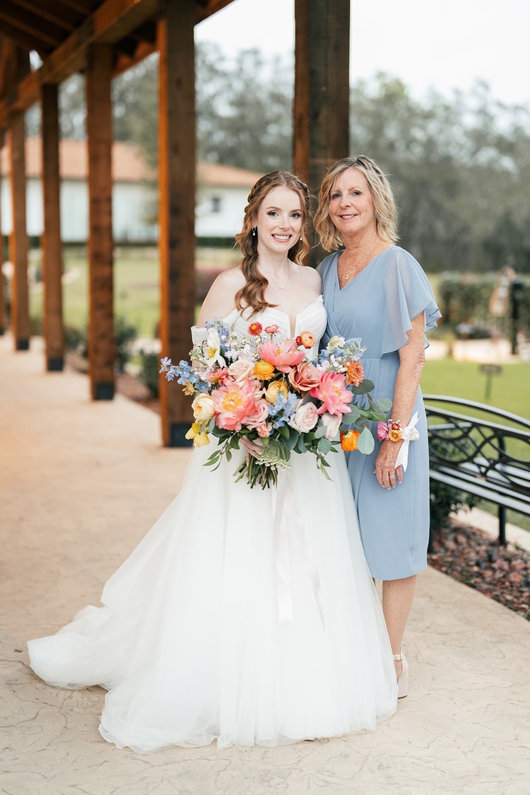 Bride smiles beside her mother under a covered walkway, holding a lush bouquet of coral, peach, and blue flowers.