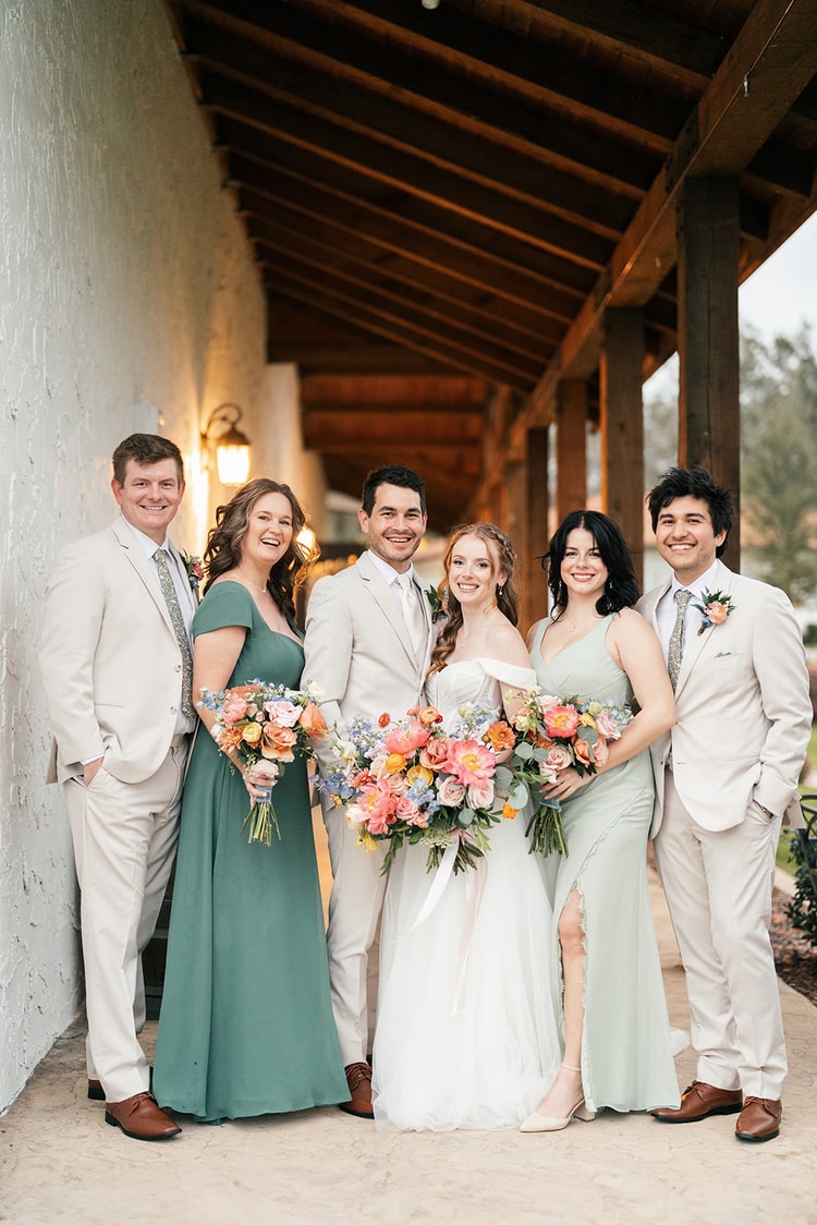 Bride and groom stand with four bridesmaids and groomsmen in sage green and tan attire under a wooden pergola, all smiling and holding bouquets.