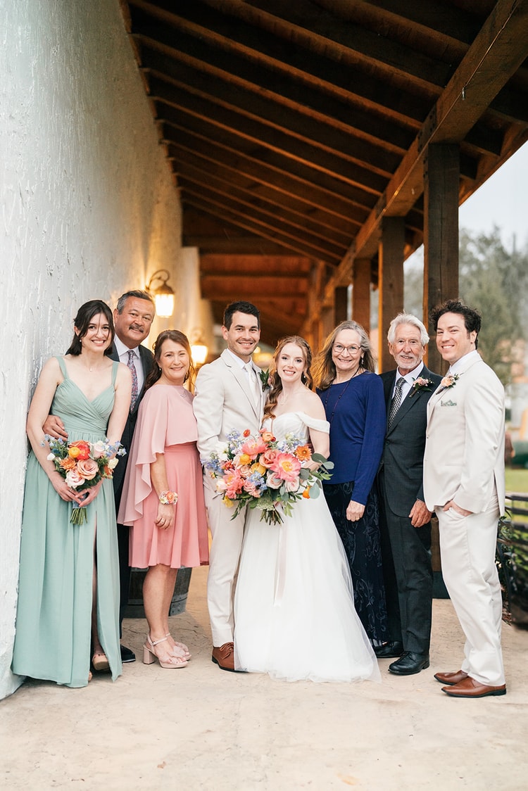 Bride and groom pose with extended family members in mixed formal attire along a stucco walkway at the wedding venue.