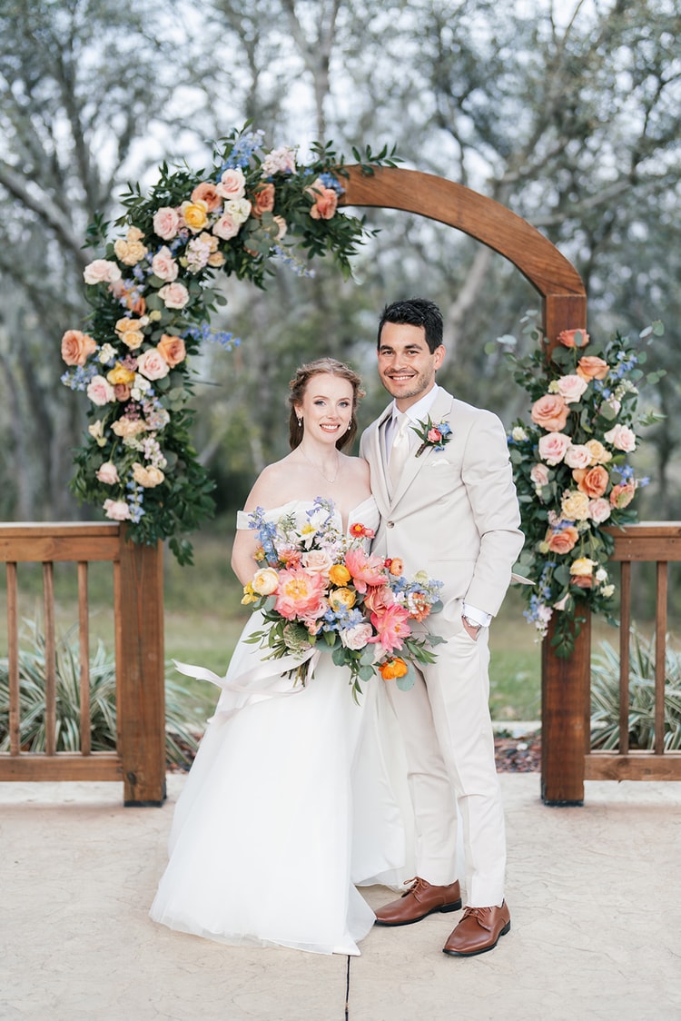 Bride and groom pose together under the floral arch, both smiling and holding her colorful bouquet.