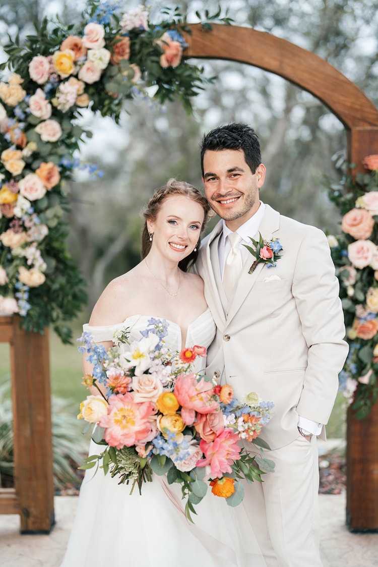 Close portrait of the bride and groom smiling at the camera under the rose and delphinium covered ceremony arch.