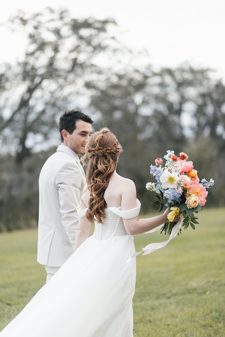 Bride, seen from behind, holds her colorful bouquet while looking back at her groom as they stand together on the lawn.