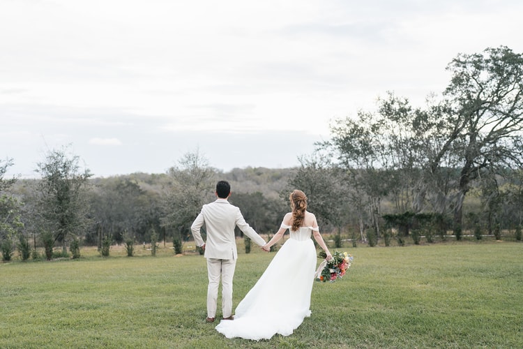 Bride and groom walk hand-in-hand away from the camera across a wide grassy field lined with mature oak trees.
