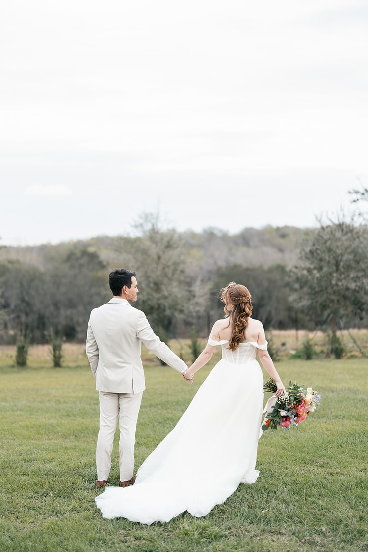 Bride and groom walk hand in hand across a grassy field, seen from behind, her bouquet of coral and blue blooms swinging at her side under an overcast sky.