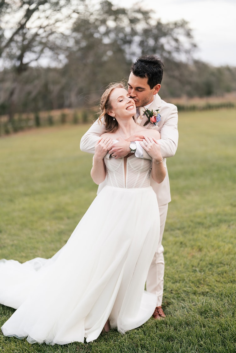 Groom kisses the bride's forehead as she laughs, arms wrapped around her from behind in an open field at Sterling Event Venue, Orlando, in warm late-day light.