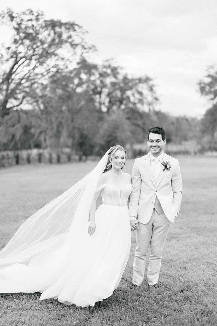 Black and white portrait of bride and groom standing hand in hand on a lawn lined with trees, both smiling toward the camera.