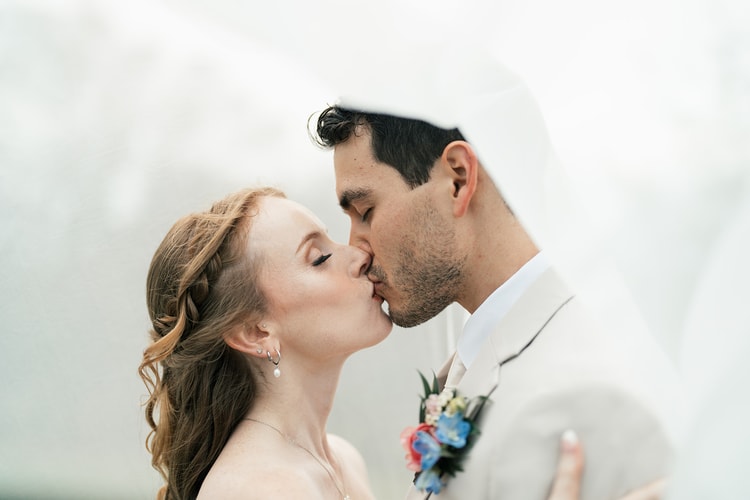 Close-up of bride and groom kissing, her braided hair and pearl earring visible, his floral boutonniere pressed between them in soft outdoor light.