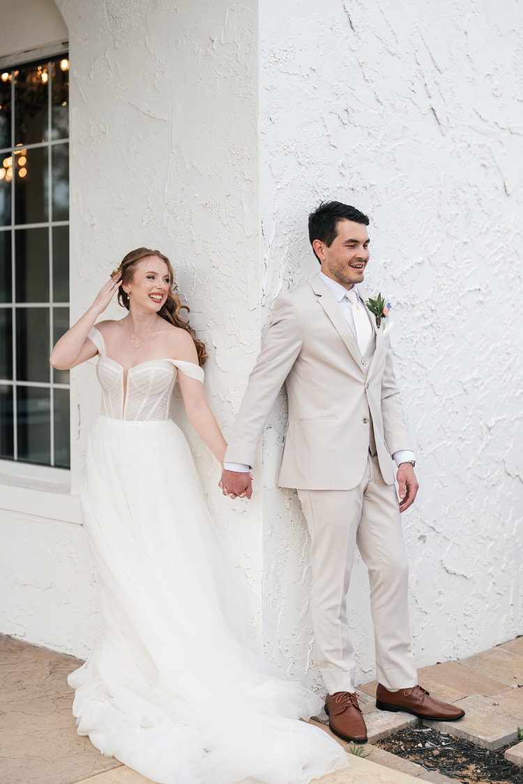Groom in a cream suit smiles while leaning against a white stucco wall outside the venue, black and white portrait.