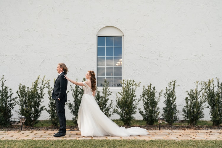 Bride looks down with hair blowing in the wind as she holds hands with the groom around a stucco corner, black and white portrait.
