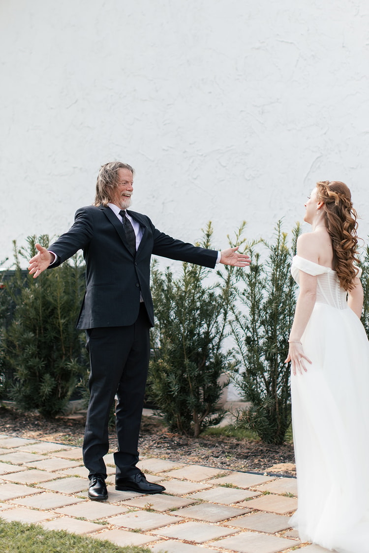 Bride's father throws his arms open with a delighted laugh as he sees her for the first time, standing on a paved path beside the venue.