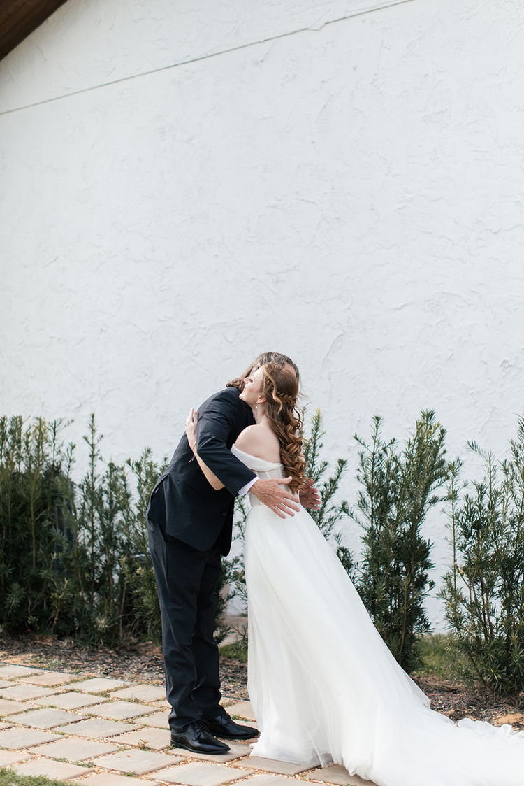 Bride's father embraces her warmly during their first-look moment, both smiling beside a hedge along the venue's exterior.