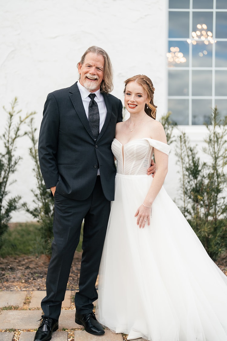 Bride and her father pose together smiling outside the venue, him in a black suit and her in her off-shoulder wedding gown.