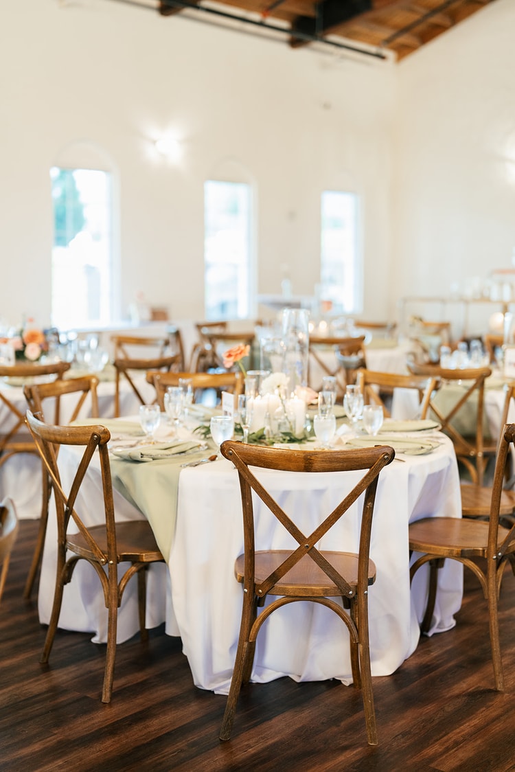 Rows of empty wooden cross-back chairs at round reception tables set with sage napkins and candlelight.