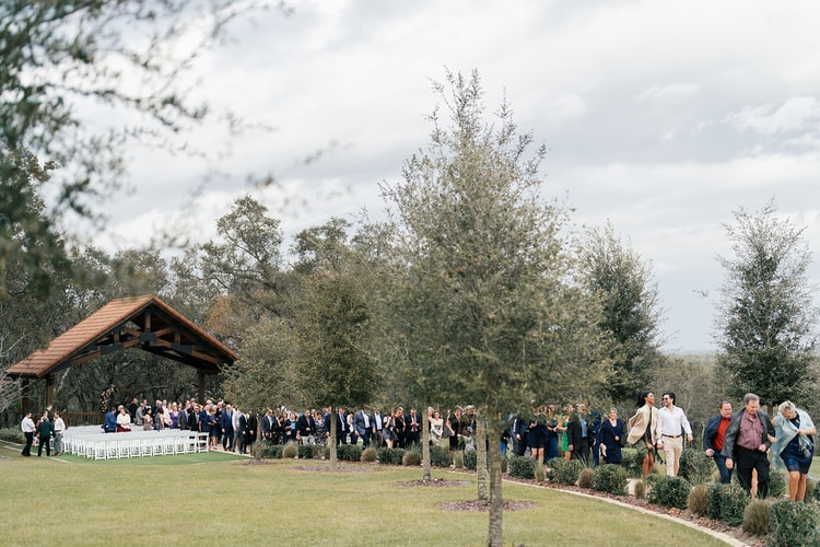 Wide shot of wedding guests walking along a landscaped path toward the reception area past rows of young oak trees.