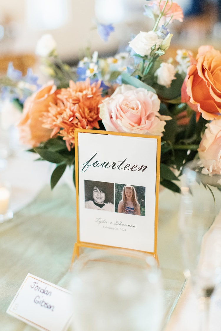 Table number sign reading 'fourteen' with childhood photos of the bride and groom, framed by orange and blush floral arrangements at the reception.