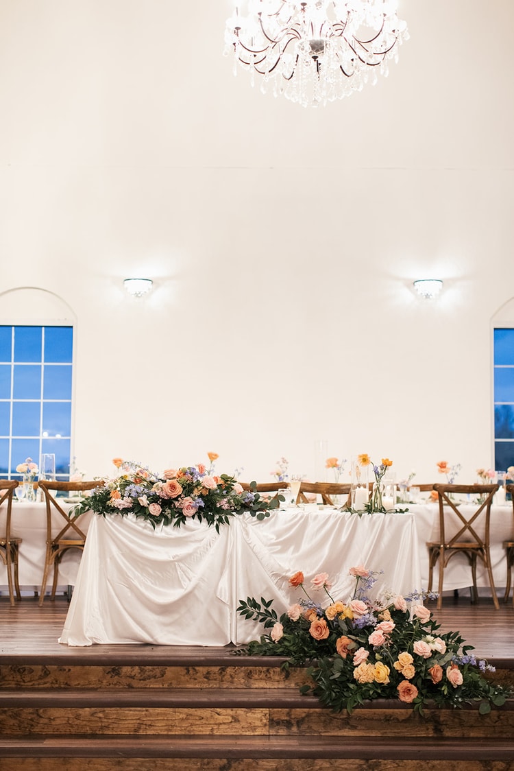 Empty reception sweetheart table draped in white linen and dressed with a garland of coral, peach, and lavender flowers, chandelier glowing overhead.