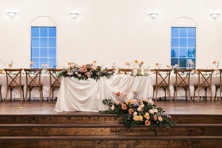 Wide view of the reception hall's sweetheart table and long guest tables set with floral garlands beneath arched windows at dusk.
