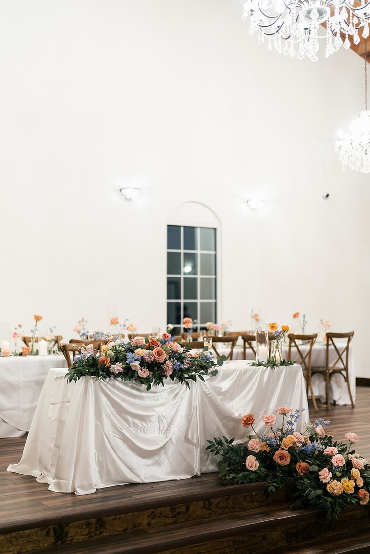 Sweetheart table on a small stage decorated with a garland of coral and lavender flowers, chandeliers hanging above an empty reception hall.