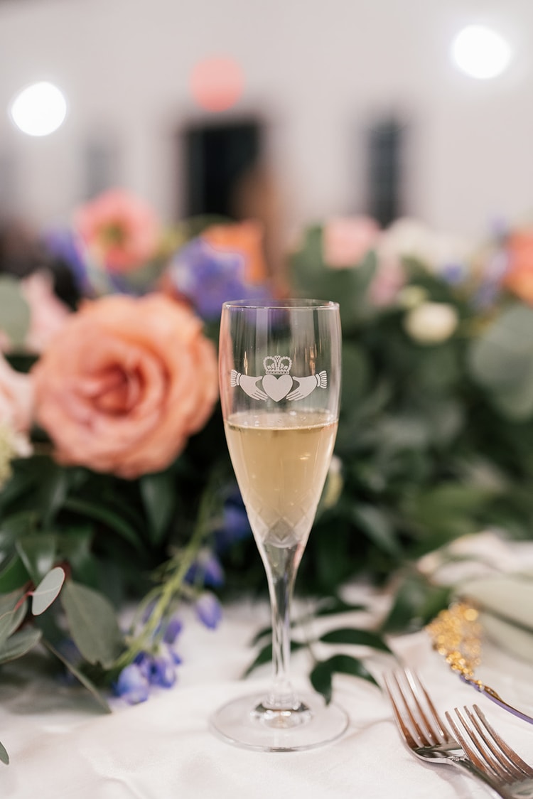 Close-up of a champagne flute etched with a Claddagh symbol, set among peach roses and greenery on the reception table.