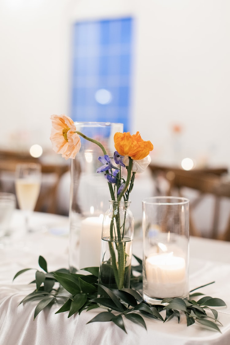 Close-up of orange poppies and purple wildflowers in a glass bud vase beside a lit candle on the reception table.